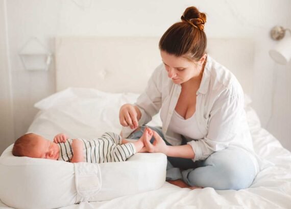 Jeune maman qui coupe les ongles de pied de son bébé en étant assise dans son lit